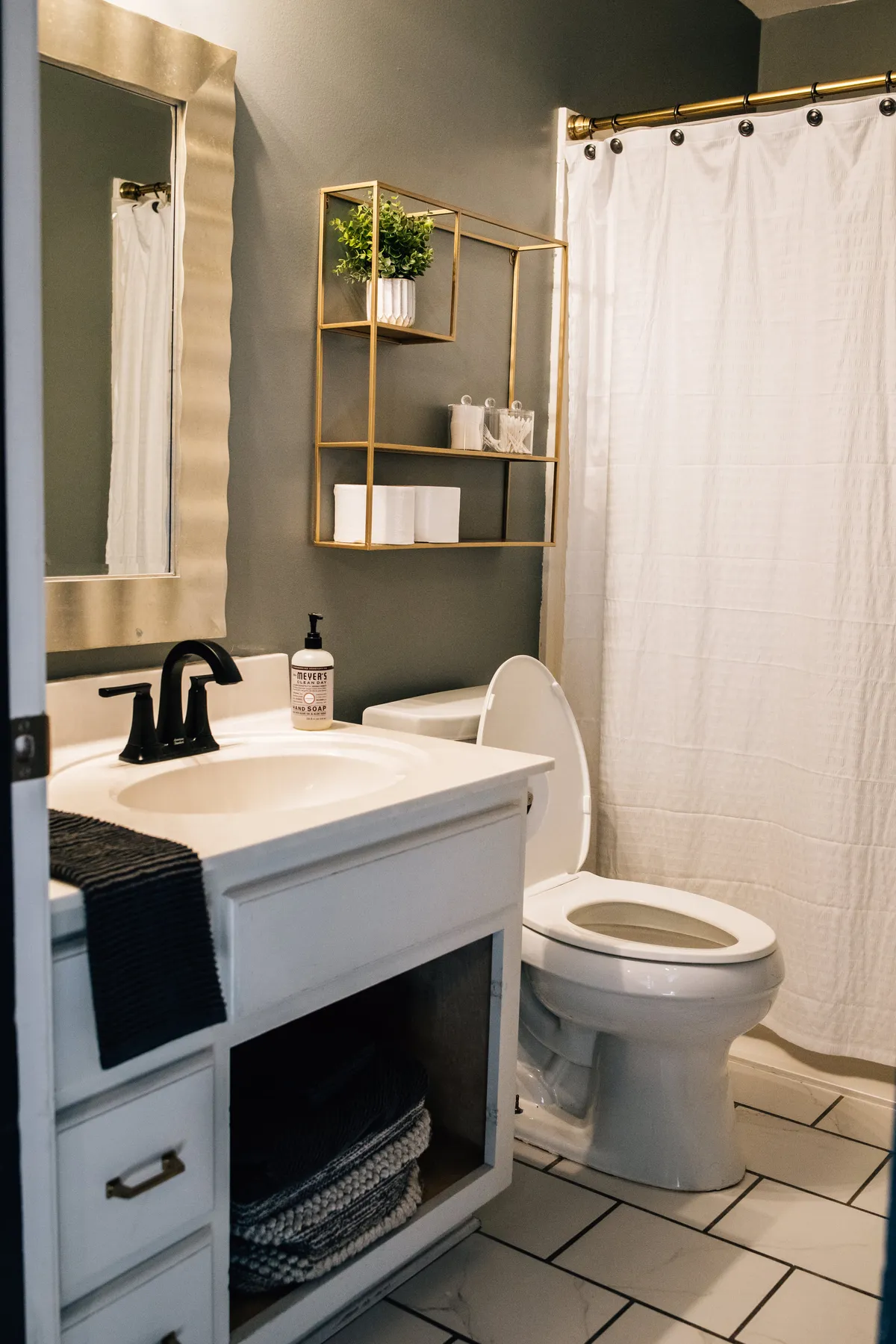 A calming bathroom space with a vanity, gold shelf, large mirror, toilet, and a shower area behind a curtain. The clean lines and elegant fixtures contribute to a sense of luxury and comfort, ideal for both solo and couples' relaxation.
