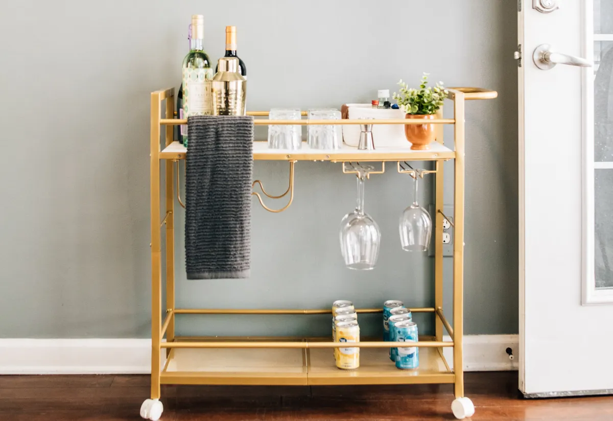 A stylish golden bar cart is set up with bottles of wine, spirits, hanging glasses, a potted plant, and colorful cans. The setup hints at a spot where guests can gather for drinks before heading out, or return to unwind after exploring the city.