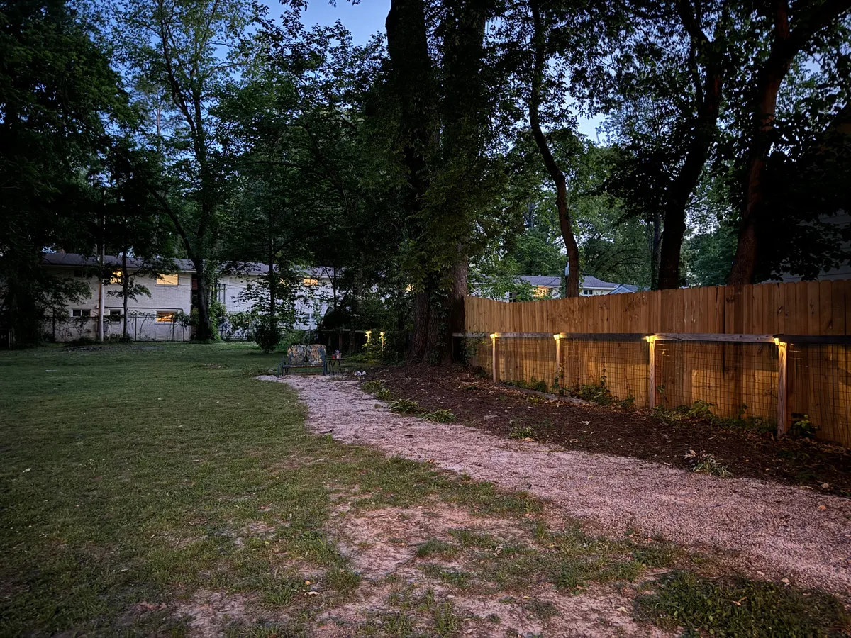 A serene exterior view of a courtyard pathway lined with lighting fixtures, trees, and a fence, suggesting a tranquil setting for evening strolls or relaxed gatherings. The moody lighting and natural elements create an atmosphere ideal for quiet reflection or intimate conversation.