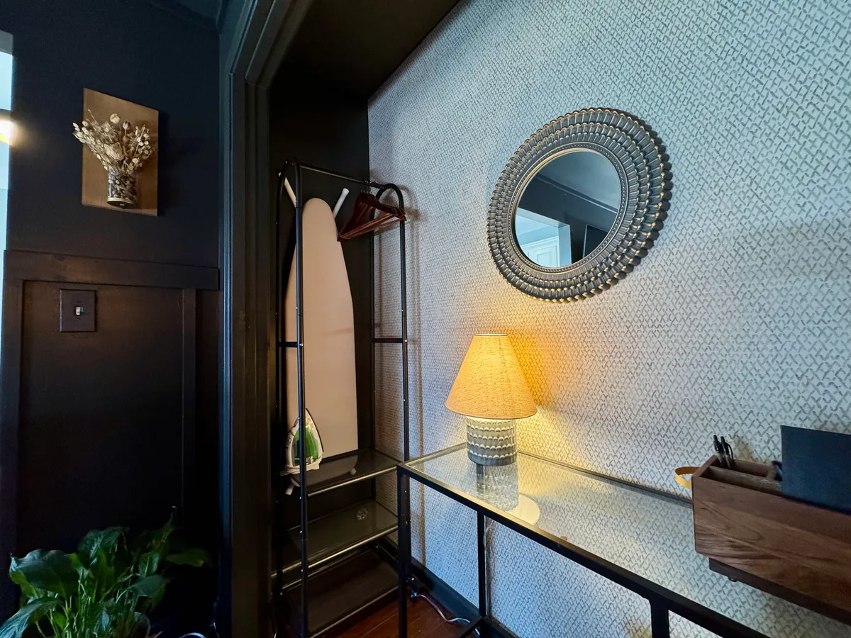 A functional and welcoming nook featuring a decorative mirror, table lamp, ironing board, potted plant, and a textured accent wall. The space conveys a sense of readiness and order, providing a glimpse into the comforts and practicalities available for business travelers.