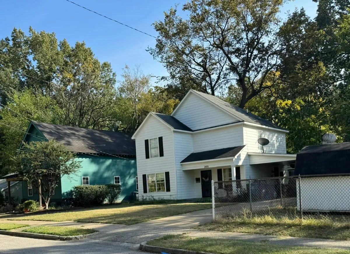 A welcoming exterior shot of a white house with black shutters and a well-kept lawn, set next to a green house. The porch and yard evoke a sense of community, relaxation, and shared outdoor moments.