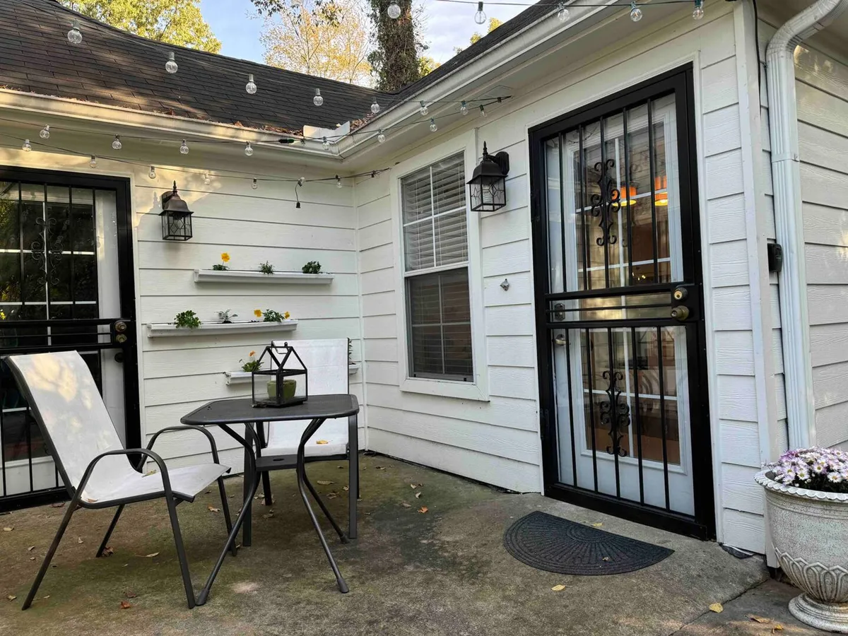 An exterior courtyard scene with a set table and chairs, decorative lanterns, vibrant flower pots, and shelving that adds a homey touch. The area is designed for outdoor dining, relaxation, and small gatherings, with greenery and cozy lighting contributing to the overall inviting feel.
