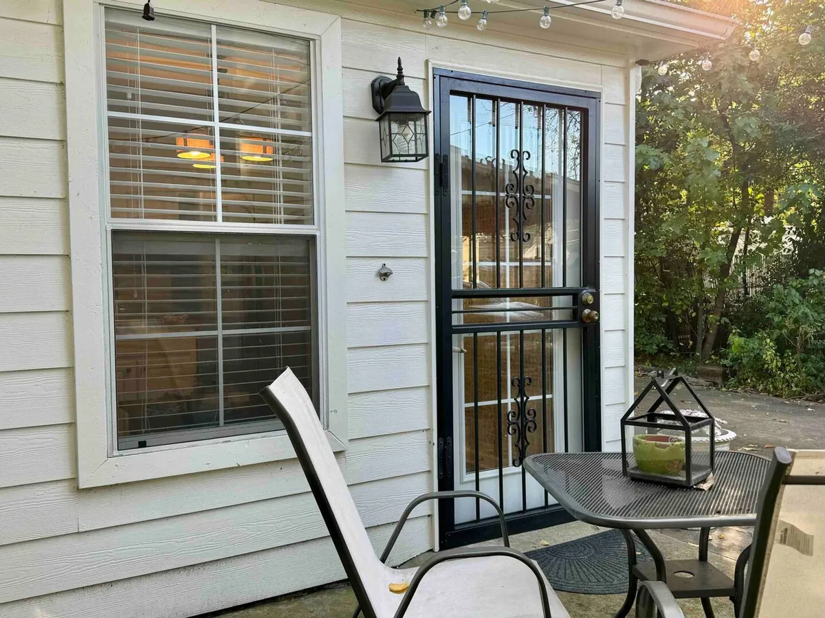 A charming exterior view featuring a decorative door framed by outdoor seating and an inviting lantern light. The space is set up for relaxation and socializing, with comfortable chairs and a welcoming ambiance that suggests both sunny daytime gatherings and cozy evening conversations under soft lighting.