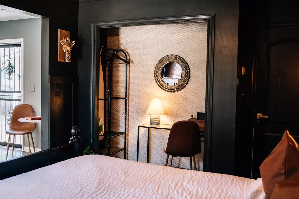 This image features a stylish studio interior with a plush bed, a modern chair, a sleek mirror, a small table, and a soft lamp casting warm ambient light. The arrangement allows for both relaxation and productive moments, reflecting a moody, inviting atmosphere.