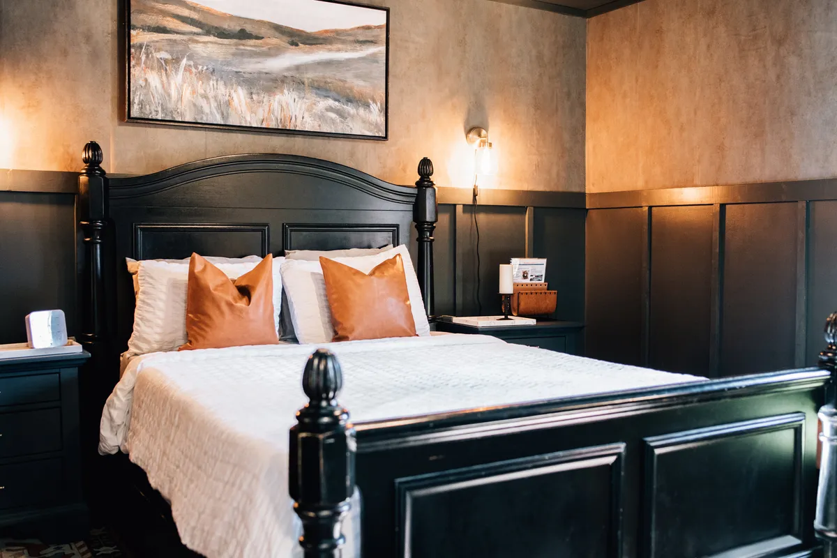 The image showcases a stylish studio interior with a bold black wooden bed frame, vibrant orange accent pillows, framed landscape artwork above the bed, and softly glowing lamps on nightstands. The space feels both moody and inviting, blending sophistication with cozy comfort.