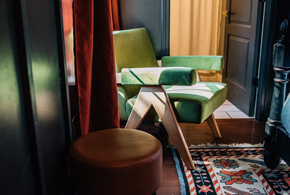 A comfortable green chair sits atop a colorful rug beside a wooden stool and light curtains, forming a serene corner for reading or unwinding. The space feels inviting and stylish, with thoughtful decor elements that reflect the apartment’s cohesive design.